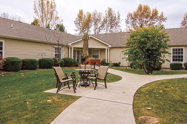 Outdoor courtyard area at Brookdale North Loveland featuring a concrete walkway, a round table with four chairs, a closed umbrella, green grass, bushes, and trees with autumn foliage surrounding a single-story beige building.