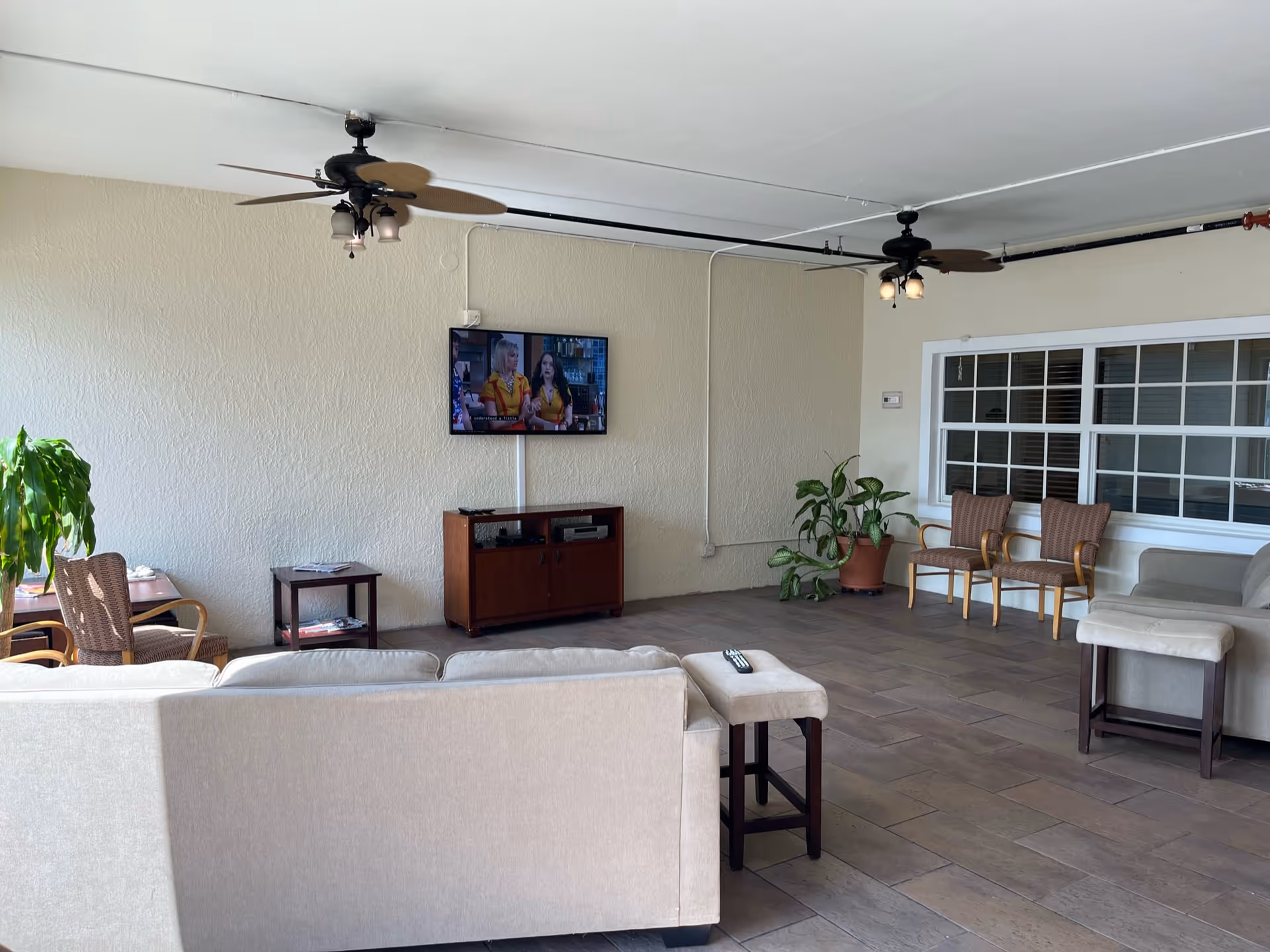 A spacious living room area with a beige sofa and matching ottoman facing a wall-mounted flat-screen TV. The room has two ceiling fans with lights, several chairs, a small wooden cabinet under the TV, and potted plants near the walls. There is a large window with white framing on the right side.