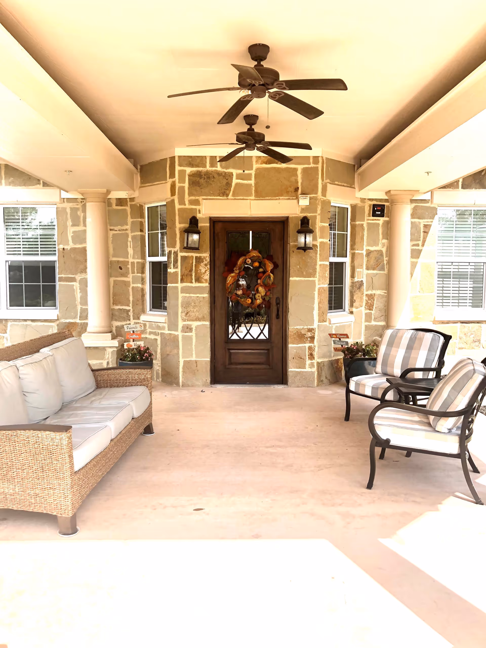 Covered outdoor patio area with a stone wall and a wooden door decorated with a fall-themed wreath. The patio has two ceiling fans, a wicker sofa with white cushions on the left, and two cushioned armchairs with striped fabric on the right. There are potted plants and decorative signs near the door.