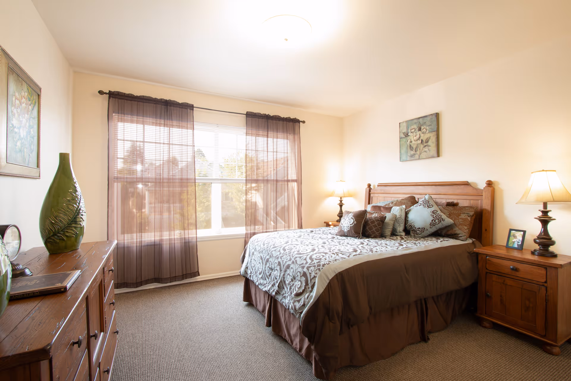 A bright and cozy bedroom with a large window covered by sheer brown curtains. The room features a wooden bed with a patterned bedspread and multiple decorative pillows. There are two wooden nightstands with lamps on either side of the bed, a wooden dresser with decorative vases, and framed artwork on the walls.