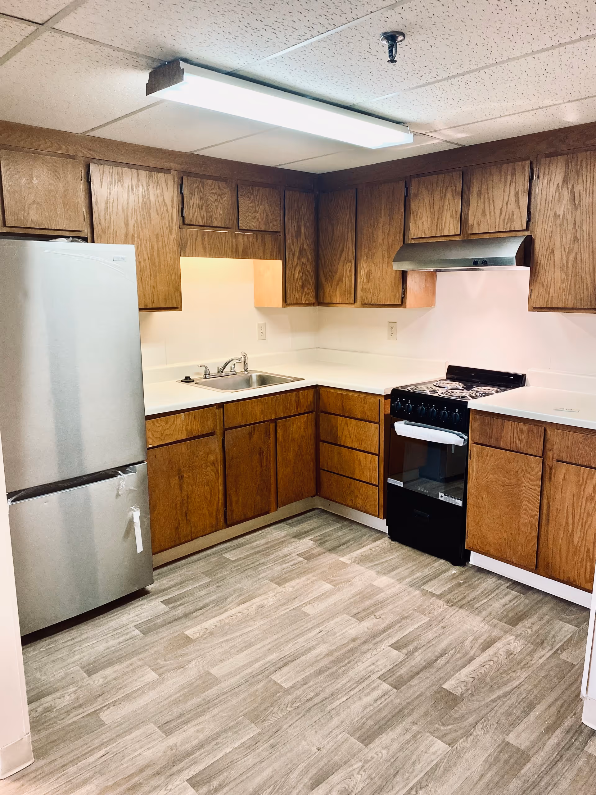 A kitchen with wooden cabinets, a stainless steel refrigerator, a black stove with an oven, a stainless steel sink, white countertops, and wood-patterned flooring under fluorescent ceiling lighting.