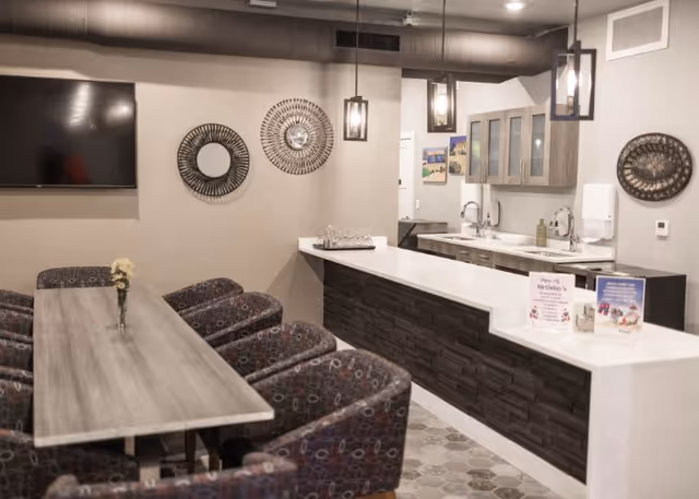 Communal dining area with a long wooden table and upholstered chairs facing a white countertop bar and kitchenette with pendant lights and wall decor.