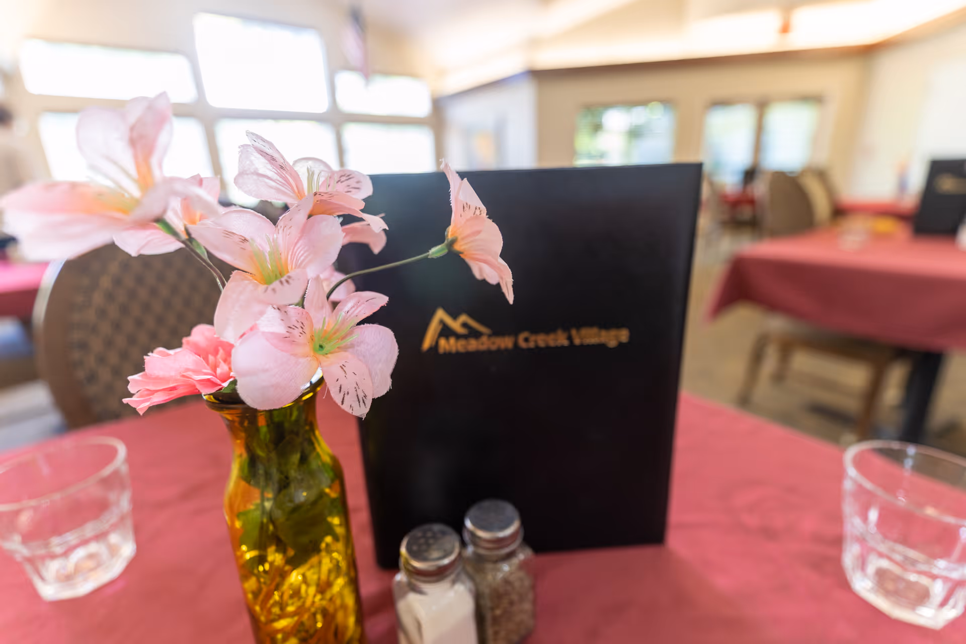 A dining table centerpiece with pink flowers in a yellow vase, salt and pepper shakers and a black Meadow Creek Village menu in a dining room.