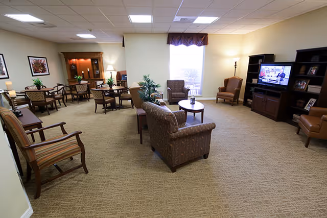 A spacious living room area in a senior living facility with multiple seating arrangements including armchairs and sofas. There is a television on a dark wooden entertainment center, a coffee table, several side tables with lamps, framed artwork on the walls, and a window with a valance letting in natural light.