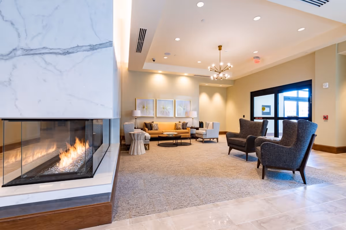 A modern and cozy living room area in a senior living facility featuring a corner glass fireplace with a marble surround, a beige sofa with decorative pillows, two armchairs, a round coffee table, side tables with lamps, and a contemporary chandelier. The room has beige walls, carpeted flooring, and large glass doors leading outside.
