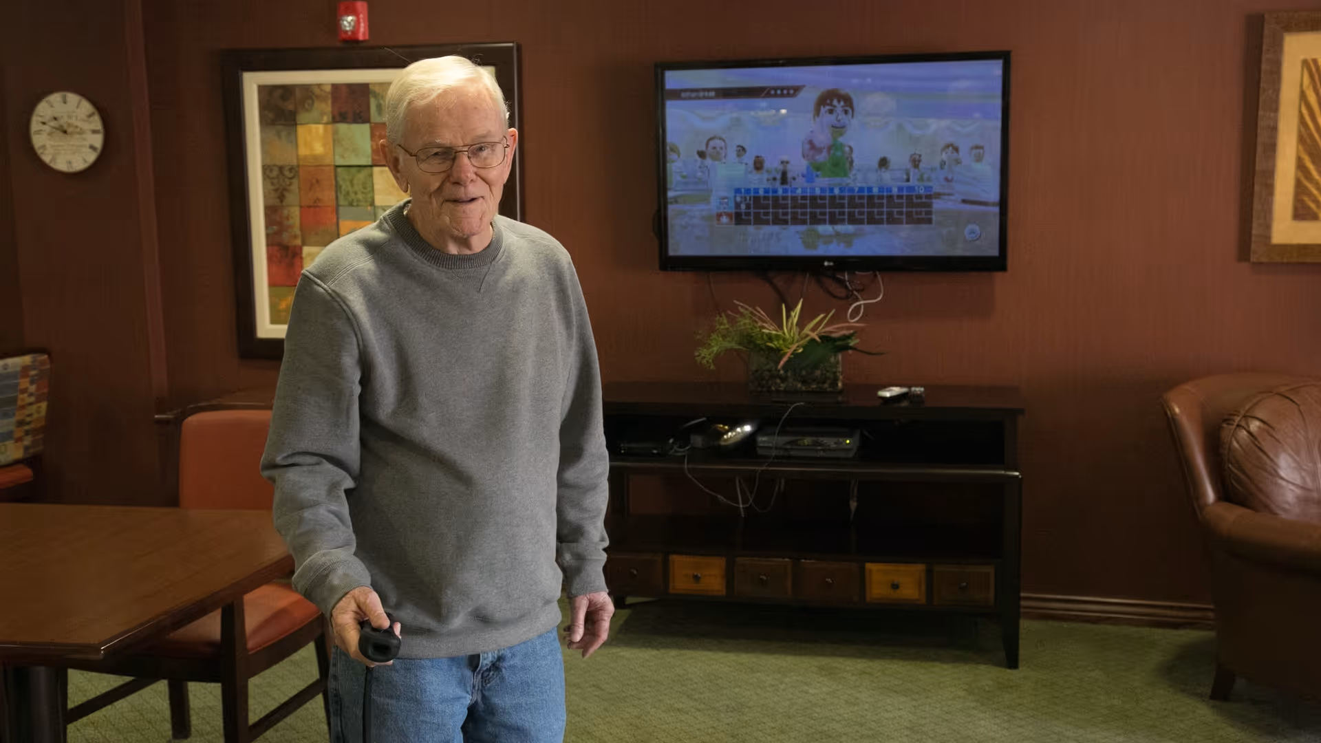 An older man stands in a lounge holding a game remote in front of a wall-mounted TV and seating.