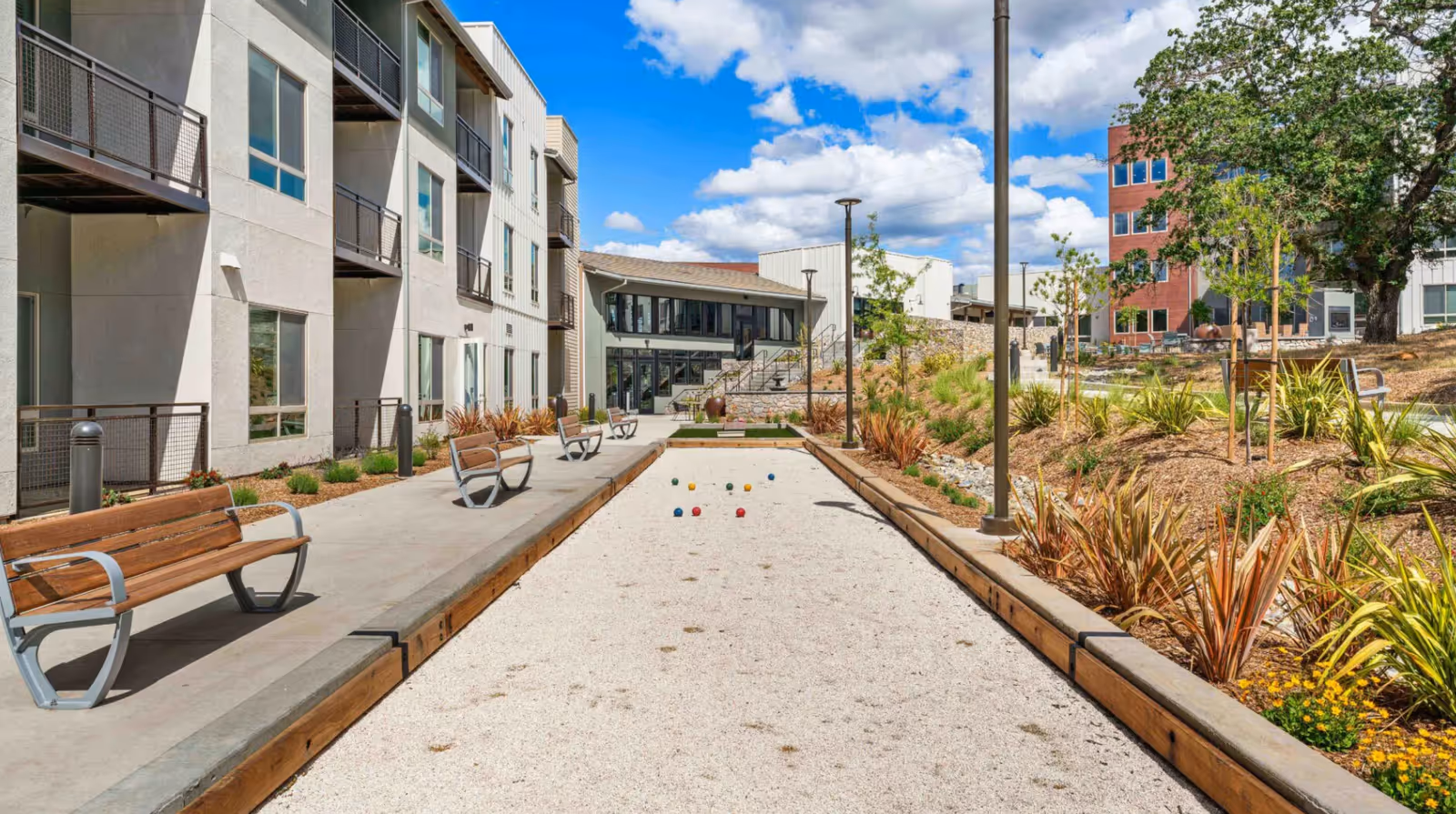 Outdoor bocce ball court with benches along the side, surrounded by landscaped plants and trees, adjacent to a multi-story building under a partly cloudy blue sky.
