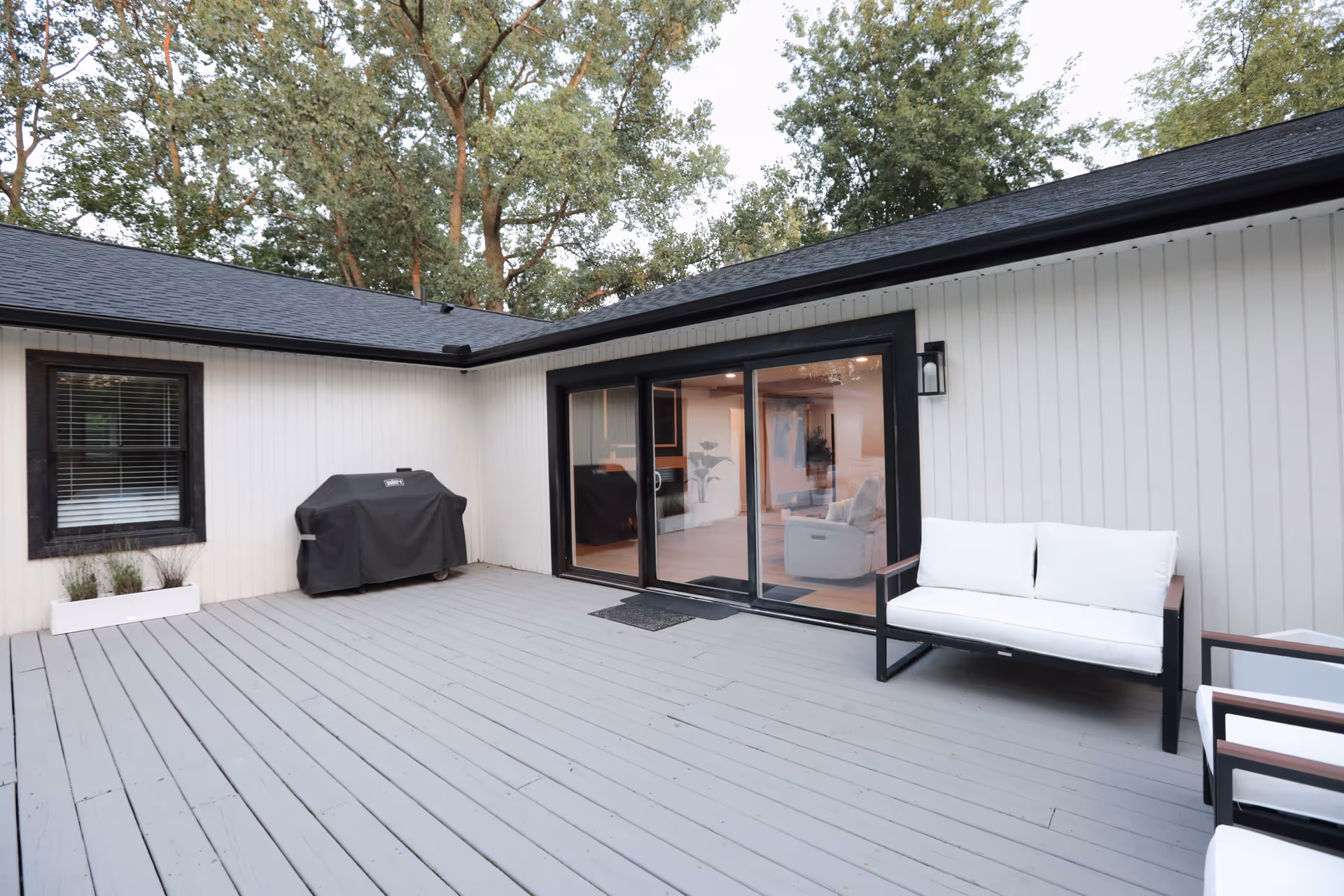 Outdoor patio area with gray wooden decking, a covered barbecue grill, white cushioned seating, and sliding glass doors leading inside a building surrounded by trees.