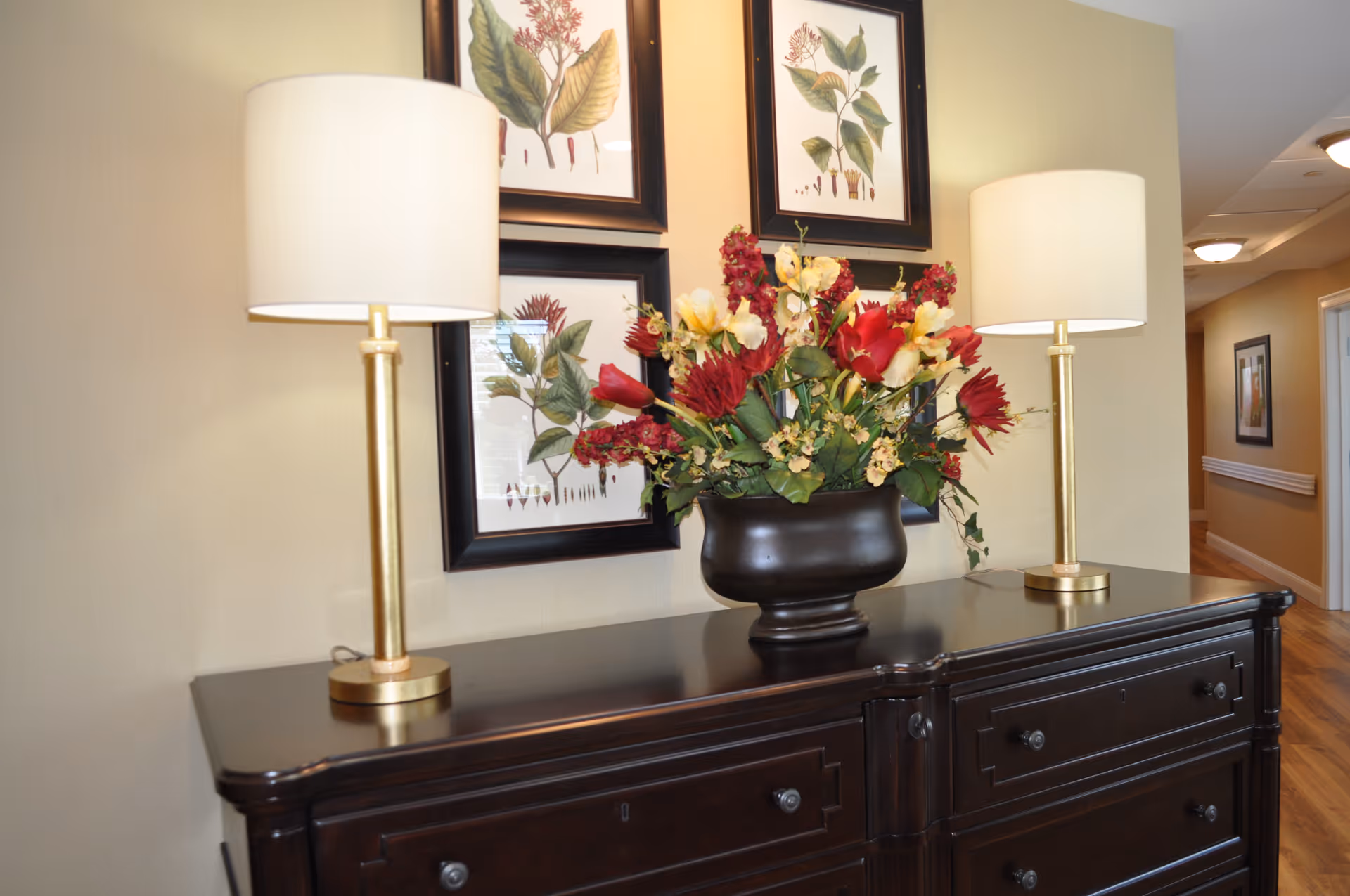 A dark wooden sideboard with two gold and white lamps on either end and a large black vase filled with red and yellow flowers in the center. Behind the sideboard, there are four framed botanical prints on a beige wall. A hallway with wooden flooring and additional framed artwork is visible to the right.