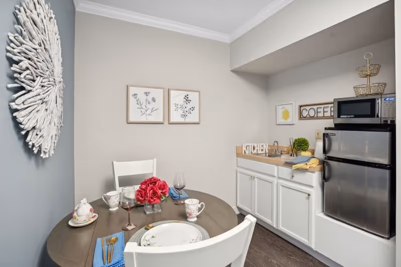 A small kitchen and dining area with a round wooden table set for two, featuring floral china, wine glasses, and a vase with red flowers. The kitchen area includes white cabinets, a countertop with a small sink, a microwave, and a mini refrigerator. Wall decorations include framed botanical prints, a decorative wooden sign that says 'COFFEE', and a white driftwood-style circular wall art.