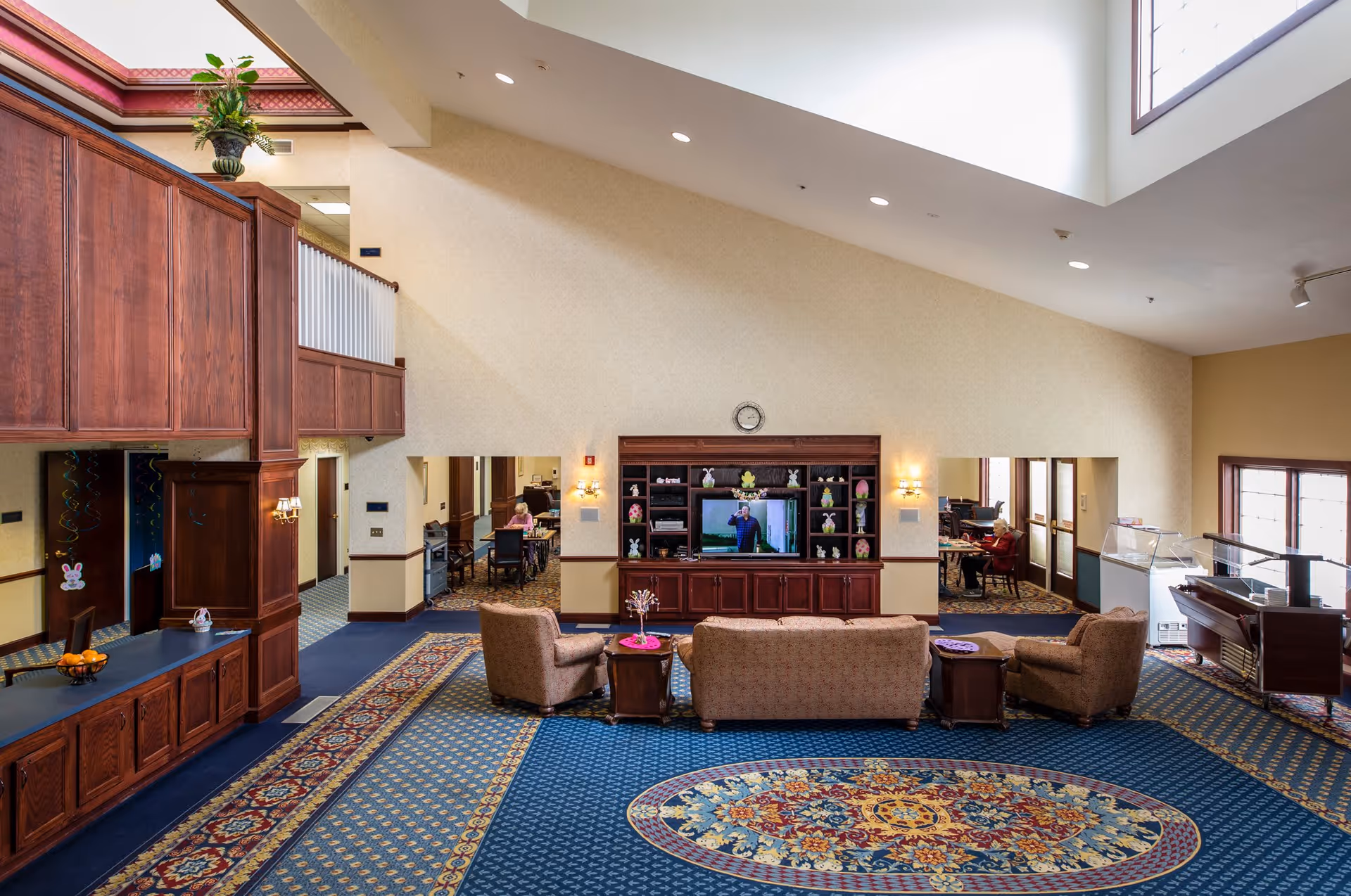 A spacious assisted living facility common area with high ceilings and large windows. The room features a patterned carpet with a central oval design, a wooden entertainment center with a television and decorative items, and seating including a sofa and two armchairs arranged around a small table. There are additional rooms visible in the background with people seated at tables. The walls are light-colored, and there is wooden cabinetry and trim throughout the space.