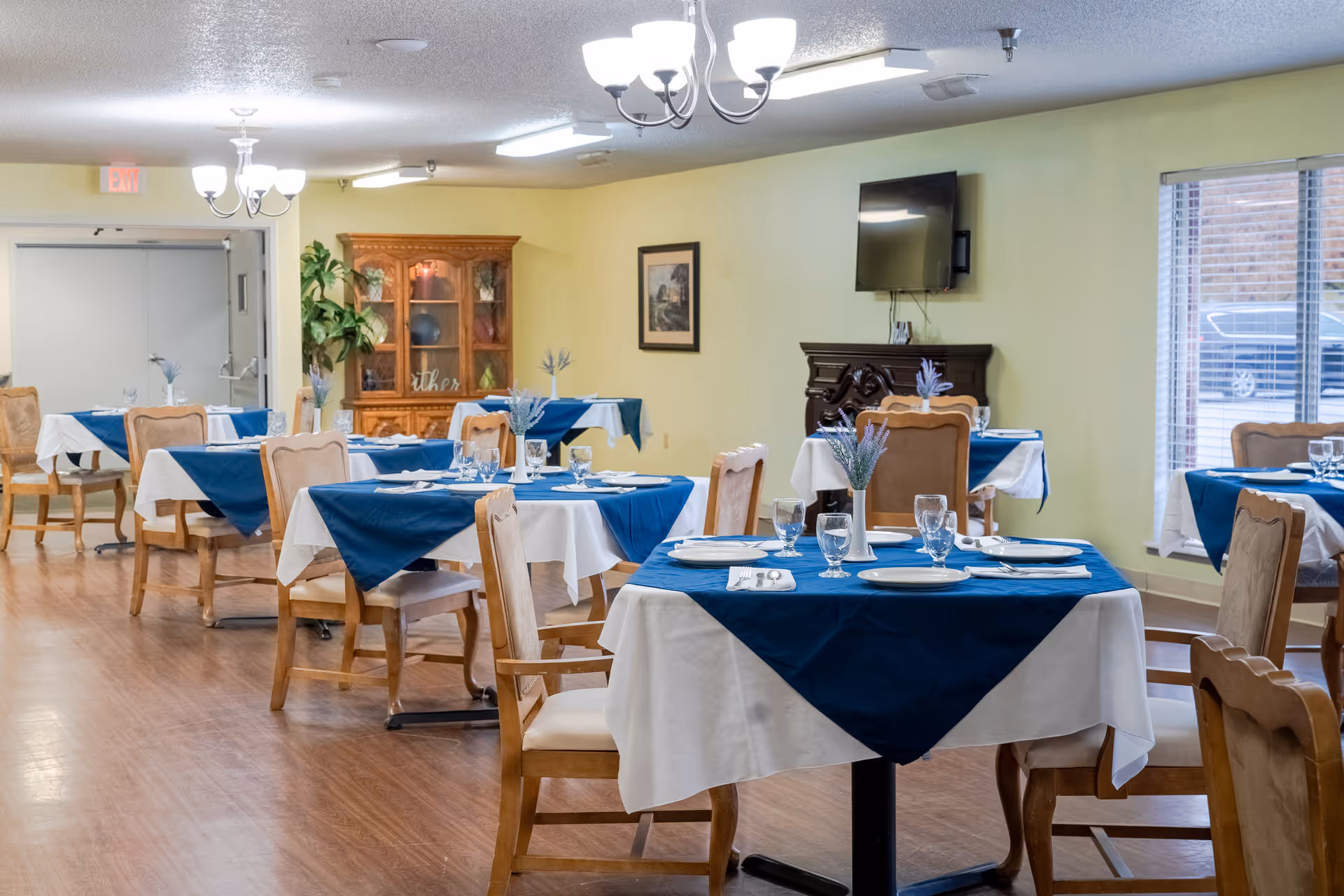A dining room with several tables covered in white tablecloths and blue table runners. Each table is set with plates, glasses, and silverware, and has a small vase with purple flowers. Wooden chairs with cushioned seats surround the tables. The room has light yellow walls, a wooden floor, a wooden cabinet with glass doors, a wall-mounted TV above a dark wooden fireplace mantel, and windows with blinds letting in natural light.