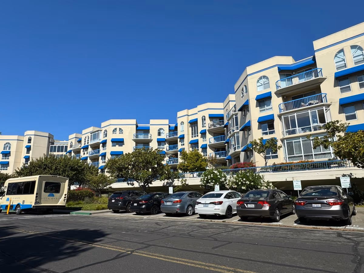 Front exterior of a multi-story beige apartment building with blue awnings and balconies and parked cars in front under a clear blue sky.