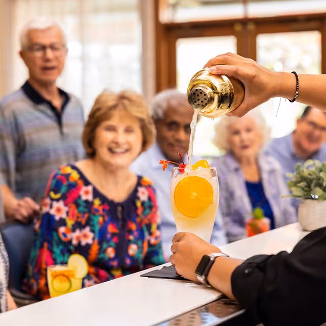 A bartender is pouring a cocktail garnished with orange slices and a cherry into a glass at a bar counter. In the background, a group of elderly people are sitting and smiling, enjoying the social atmosphere inside a senior living facility.