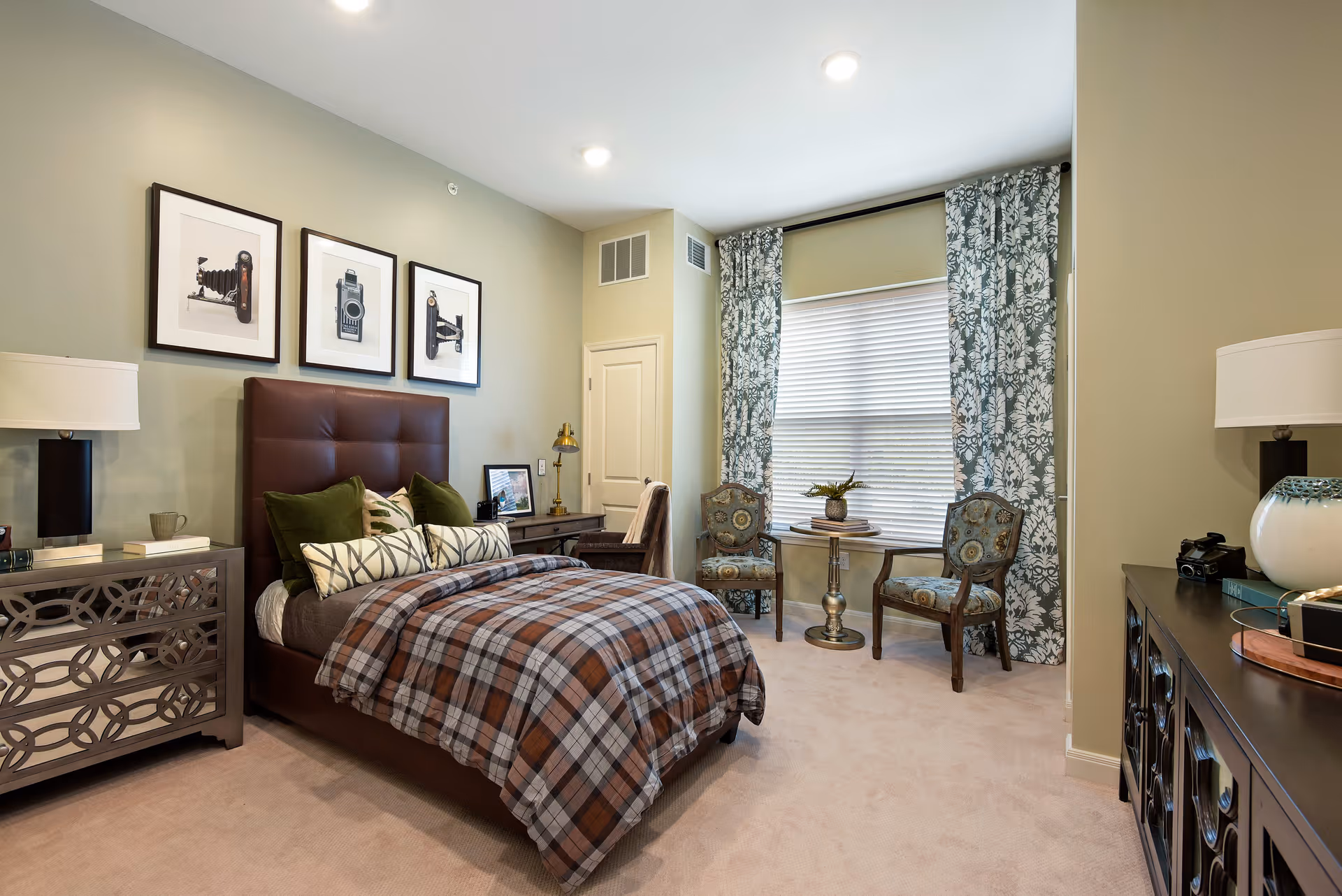 A cozy bedroom with a brown upholstered bed featuring plaid bedding and multiple pillows. On the left side, there is a mirrored nightstand with a lamp and books. Above the bed, three framed pictures of vintage cameras are hung on the wall. To the right, there is a window with floral curtains, two patterned chairs, and a small round table with a plant and books. A desk with a chair and lamp is positioned near the corner, and a dark wooden dresser with a lamp and decorative items is on the right side of the room.