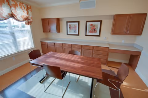 A small dining or meeting room with a rectangular wooden table surrounded by four modern chairs. The room has built-in wooden cabinets along one wall, two framed floral artworks above the cabinets, and a window with blinds and a floral valance letting in natural light.