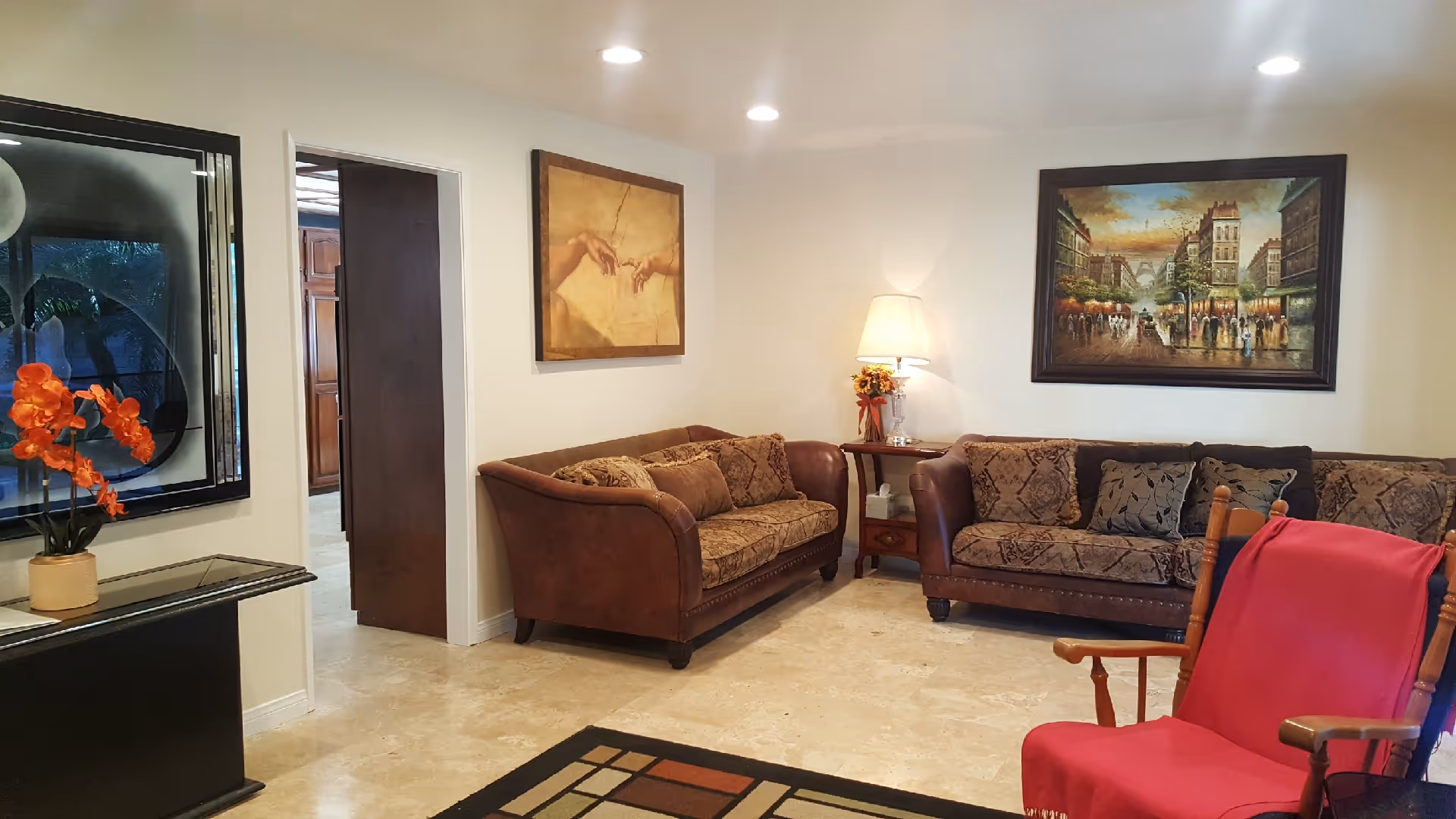A cozy living room with two brown patterned sofas, a wooden rocking chair draped with a red blanket, a side table with a lamp and flower arrangement, and three framed paintings on the walls. The floor is tiled and there is a decorative rug in the foreground.