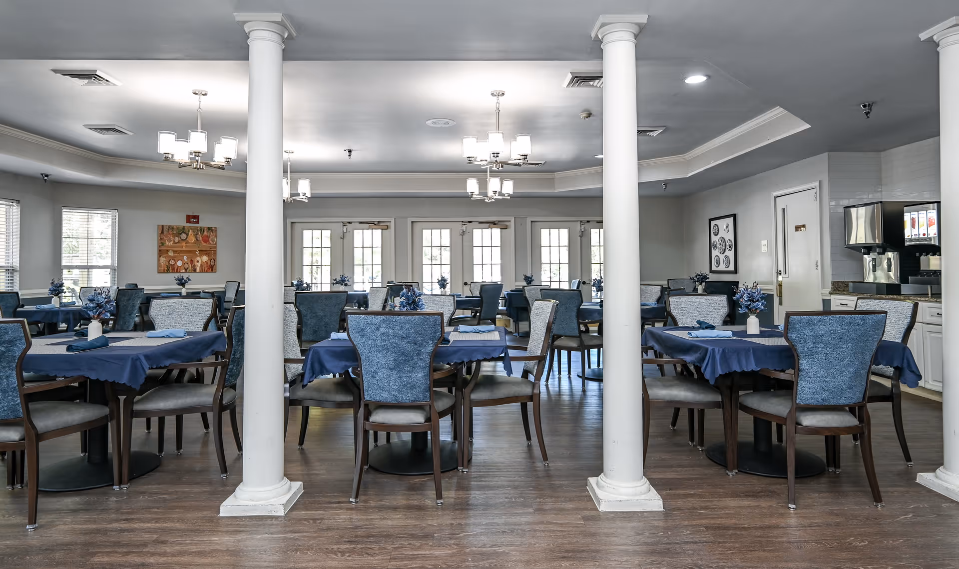 Dining room with multiple round tables set with blue tablecloths and matching upholstered chairs, white columns, chandeliers, and a beverage station.