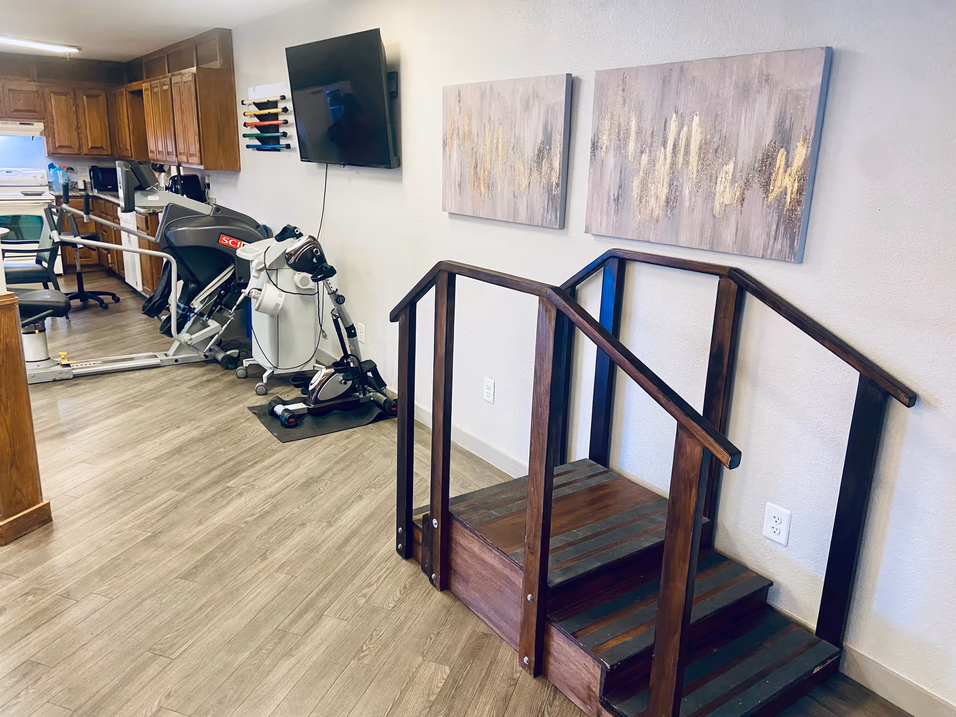 Room with exercise equipment, a wooden practice stair with handrails, wall art and a kitchenette in the background.