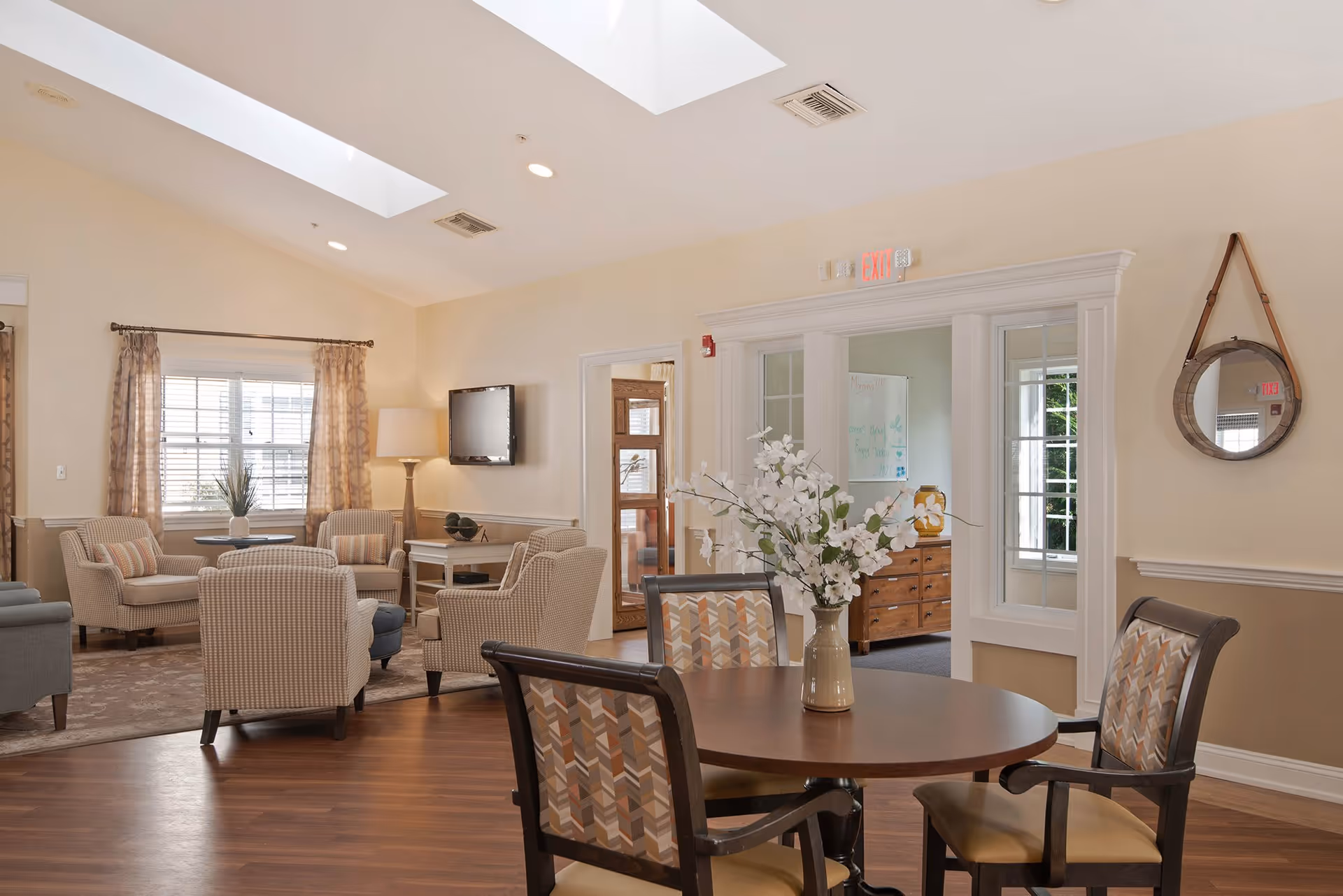 A bright and cozy common area in a senior living facility with a round wooden table and four patterned chairs in the foreground. In the background, there are several upholstered armchairs arranged around a small table near a window with curtains. A flat-screen TV is mounted on the wall, and a floor lamp stands beside the chairs. The room has light-colored walls, wooden flooring, skylights, and a round mirror hanging on the wall.