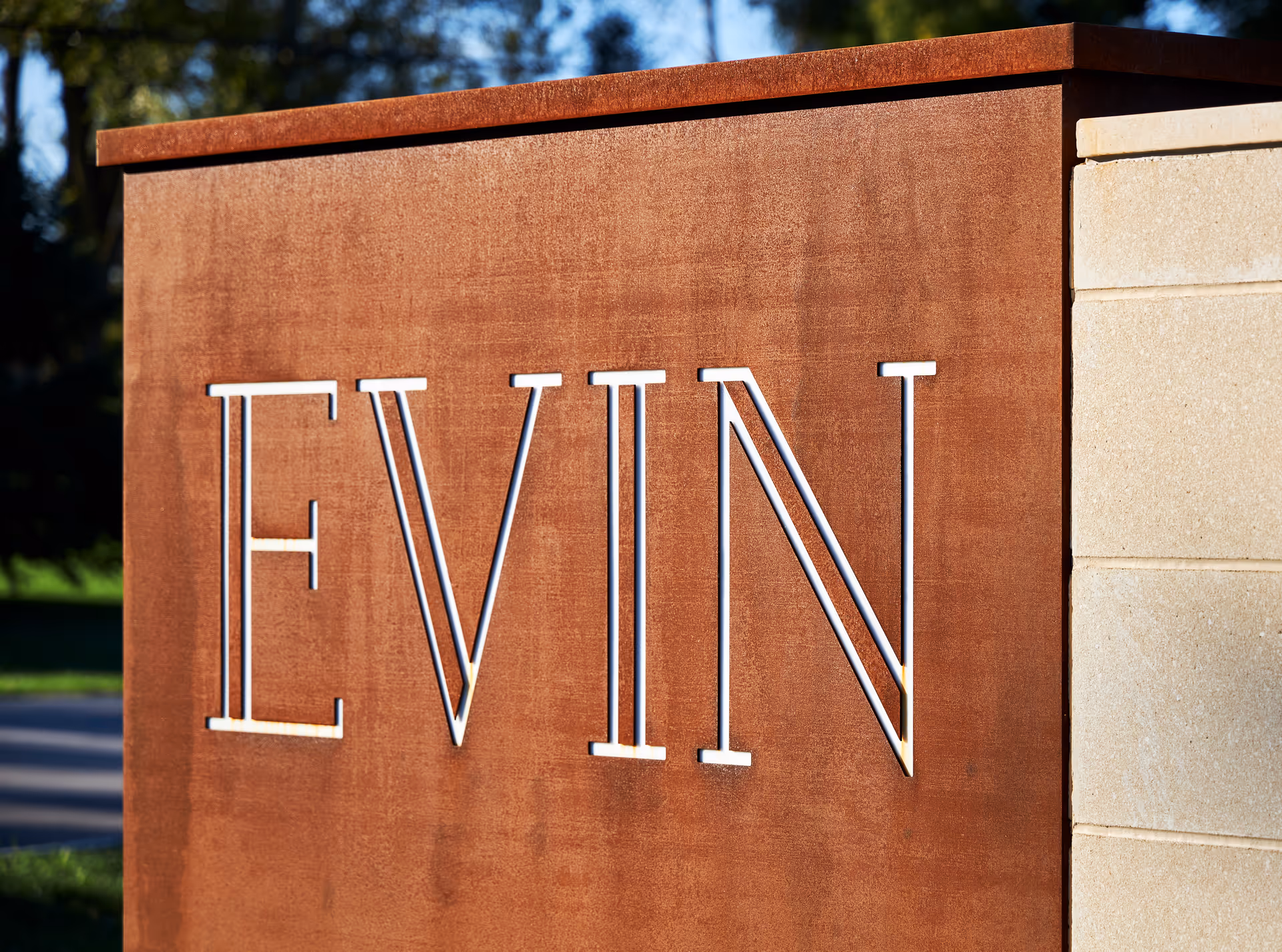Close-up of a rust-colored metal sign with the word 'EVIN' cut out in large capital letters, mounted on a stone base with greenery and trees visible in the background.