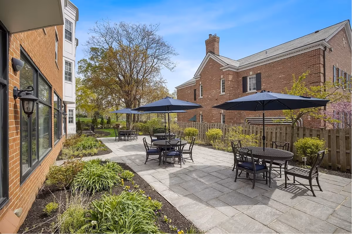 Outdoor patio area with several round tables and chairs under large blue umbrellas, surrounded by greenery and adjacent to a brick building and a wooden fence.