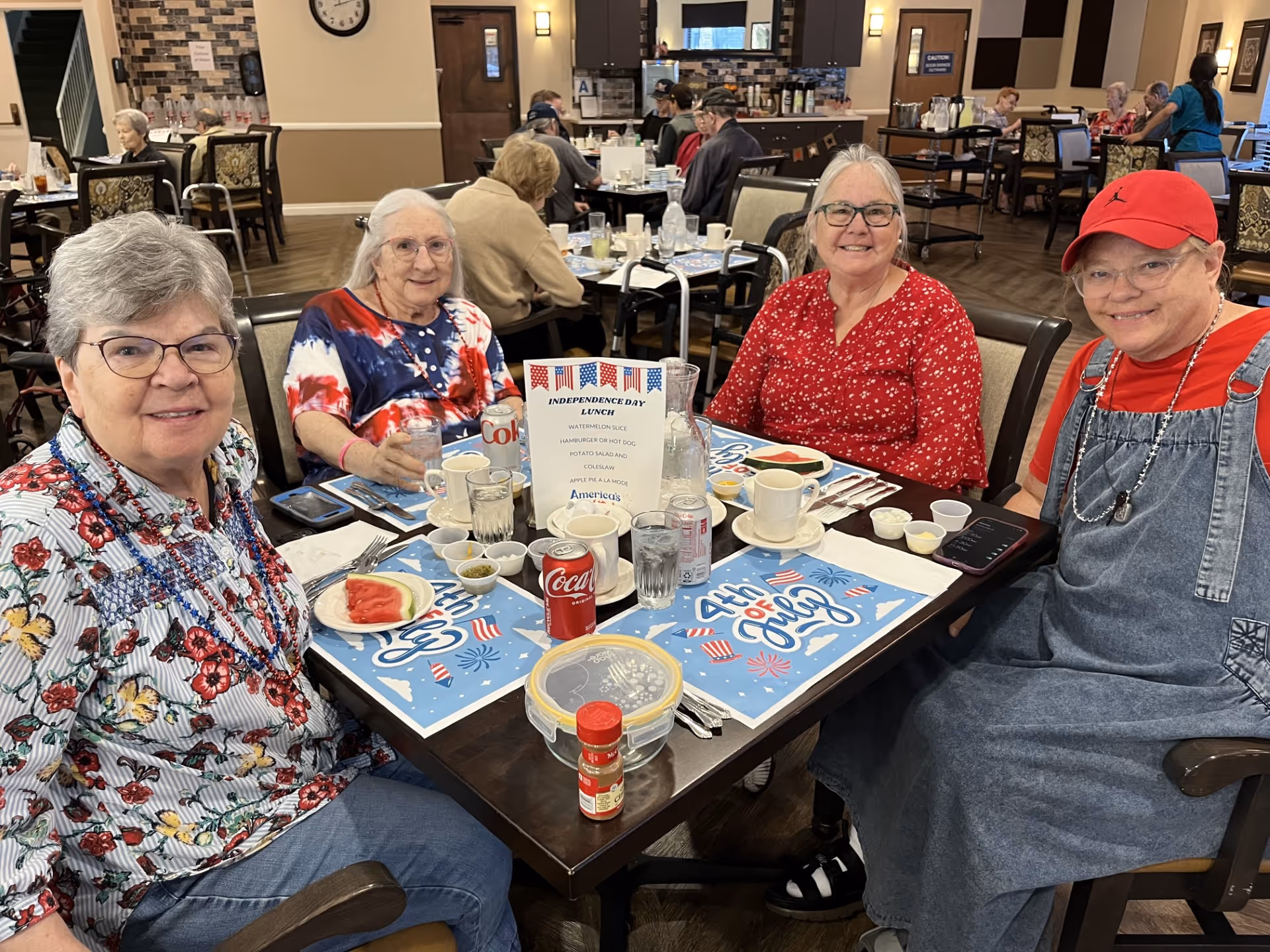Four elderly women sitting around a dining table in a senior living facility, smiling at the camera. The table is set with plates, cups, soda cans, and a menu card for an Independence Day lunch. Other residents are visible dining in the background in a well-lit dining room with wooden floors and modern decor.