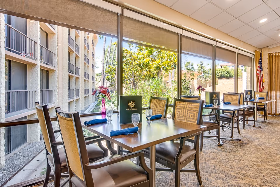 A bright dining room with several tables and chairs arranged neatly. Each table has a menu, a glass, and a blue napkin. Large windows let in natural light and offer a view of greenery and the exterior of the building. An American flag is visible in the corner of the room.