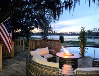 Outdoor wooden deck area with a curved wicker seating arrangement around a fire pit table. An American flag is displayed on the left side, and there is a view of a lake with trees and a sunset in the background.