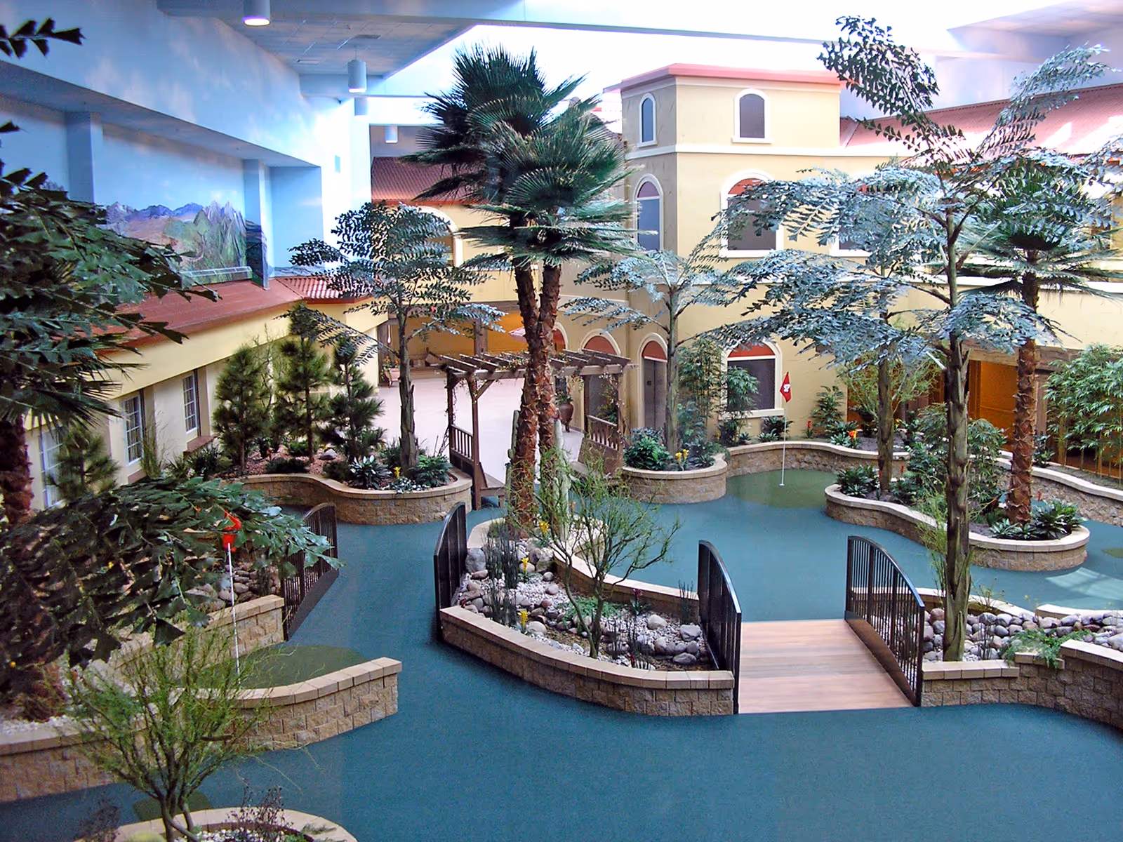 Indoor garden area with various trees and plants, a small wooden bridge over a dry rock bed, and a miniature putting green with a flag. The space is surrounded by walls painted to resemble a building facade with windows and a mural of mountains in the background.