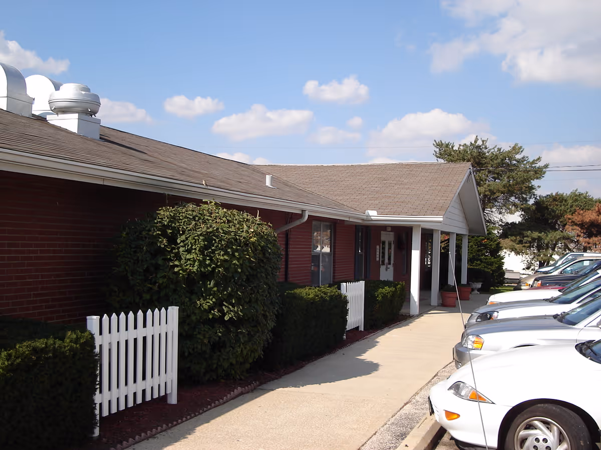Exterior view of a single-story brick building with a sloped roof, white trim, and a small covered entrance. There are bushes and a white picket fence along the sidewalk leading to the entrance. Several cars are parked in a row along the right side of the building under a partly cloudy sky.