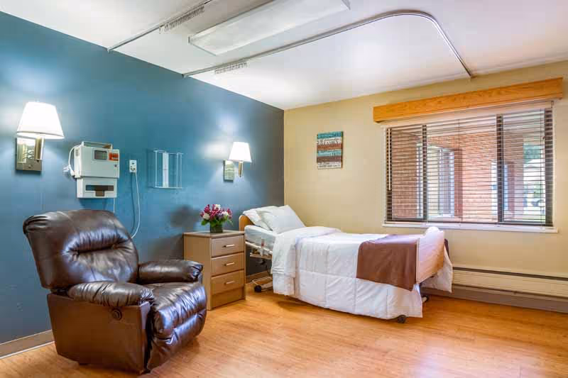 A bright senior care bedroom with a single bed, bedside table, leather recliner, wall lamps and a window with blinds.