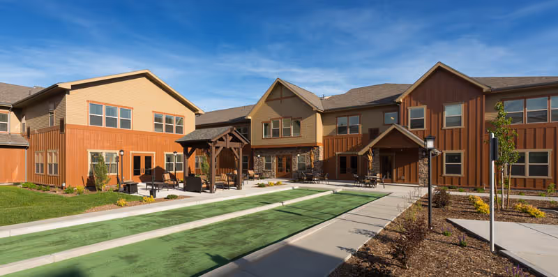 Exterior courtyard of a senior living community showing two-story wood-paneled buildings, a covered seating gazebo, and green lawn areas under a clear blue sky.
