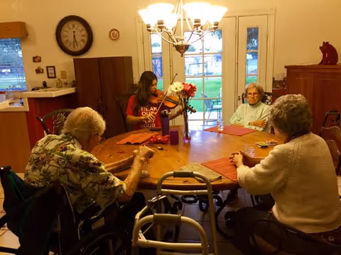 Three elderly women sitting around a wooden dining table in a well-lit room, with one woman playing a violin. There is a walker near the table, a clock on the wall, and a glass door leading outside in the background.