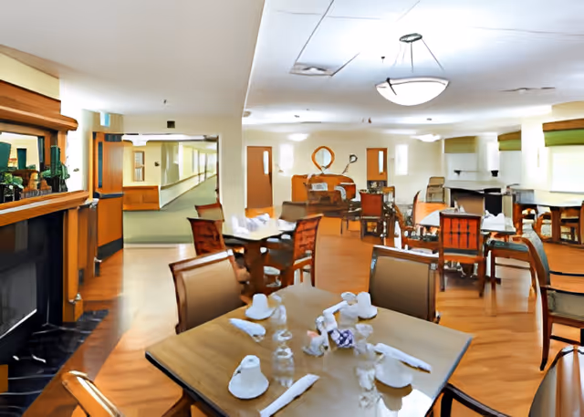 Spacious dining room in a senior living facility with multiple set tables and wooden chairs on a hardwood floor.