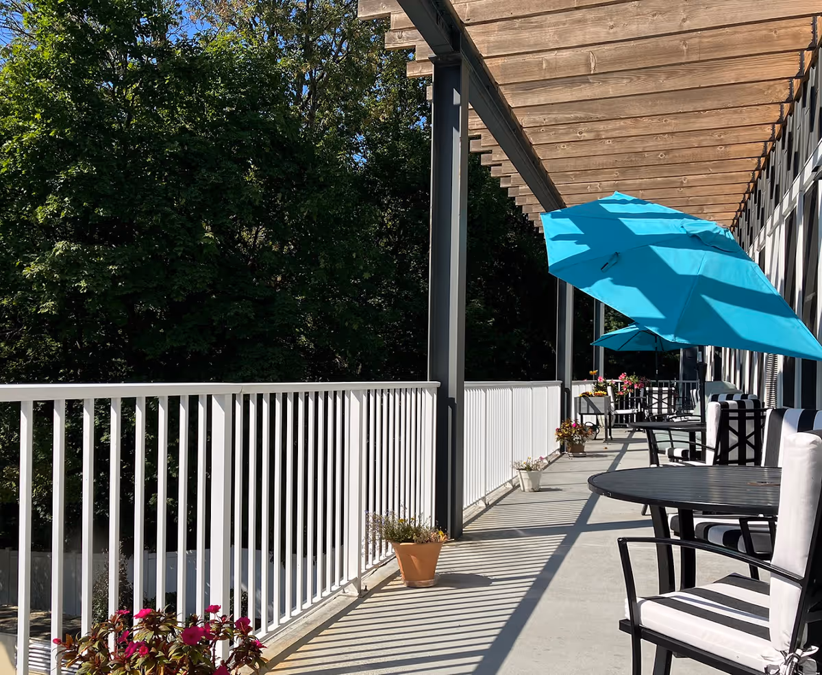 Outdoor patio area with white railing, black metal tables and chairs with white cushions, blue umbrellas providing shade, and potted plants along the walkway. Trees and greenery are visible in the background.