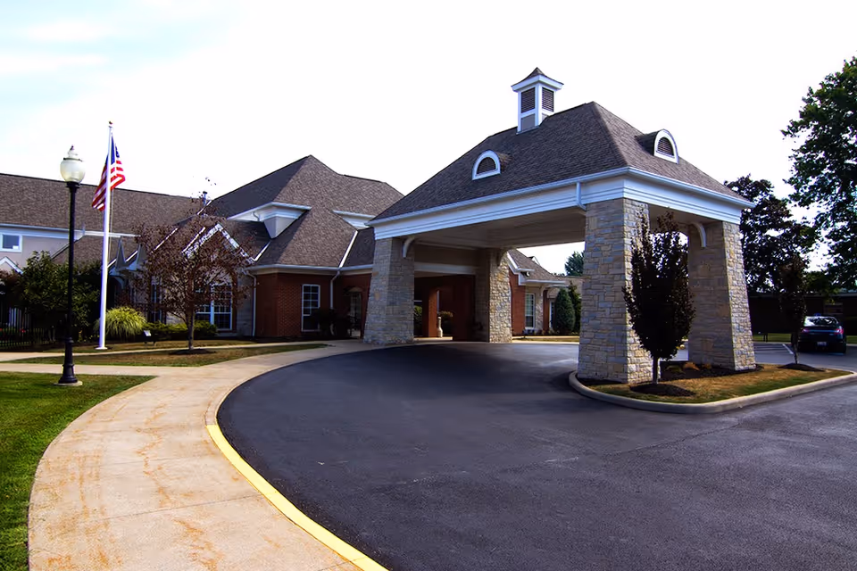Covered porte-cochère entrance of a brick senior living building with a circular driveway and an American flagpole.