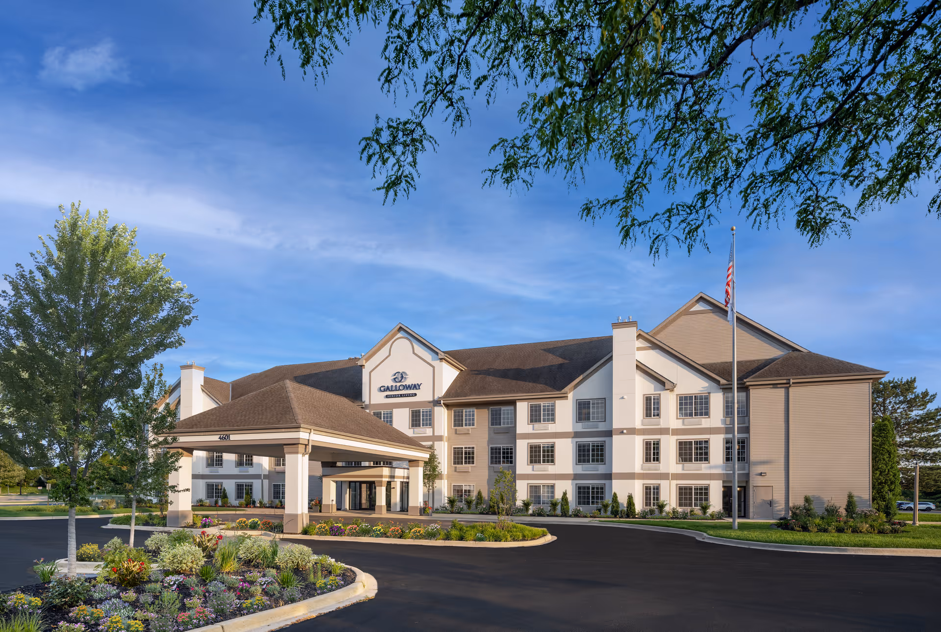 Front exterior view of Galloway Senior Living building with a covered entrance, landscaped flower beds, trees, and an American flag on a flagpole under a blue sky.