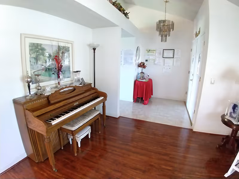 Interior entry/living area showing an upright piano against a wall, hardwood floors, and a small table with a red cloth in the tiled foyer.
