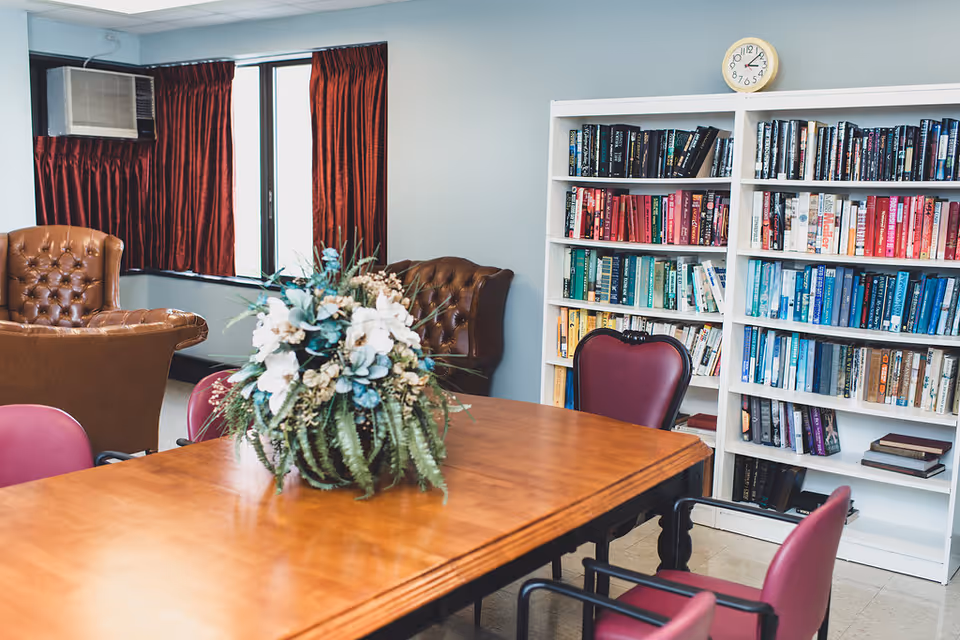 A cozy room with a wooden table surrounded by maroon chairs, a floral centerpiece on the table, two brown leather armchairs near a window with red curtains, a white bookshelf filled with books organized by color, and a clock on the wall above the bookshelf.
