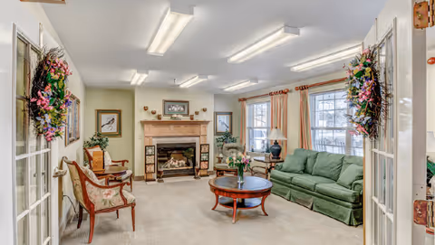 Communal living room with a green sofa, fireplace, round coffee table, armchairs, and large windows.