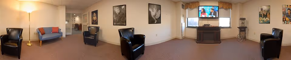 A spacious living room area with beige walls and carpeted floor. The room features three black leather armchairs and a blue loveseat with orange pillows. There is a tall floor lamp on the left side and several framed black-and-white portraits on the walls. A flat-screen TV is mounted on the wall above a dark wooden cabinet, with two windows on either side. A popcorn machine is visible near the right window.