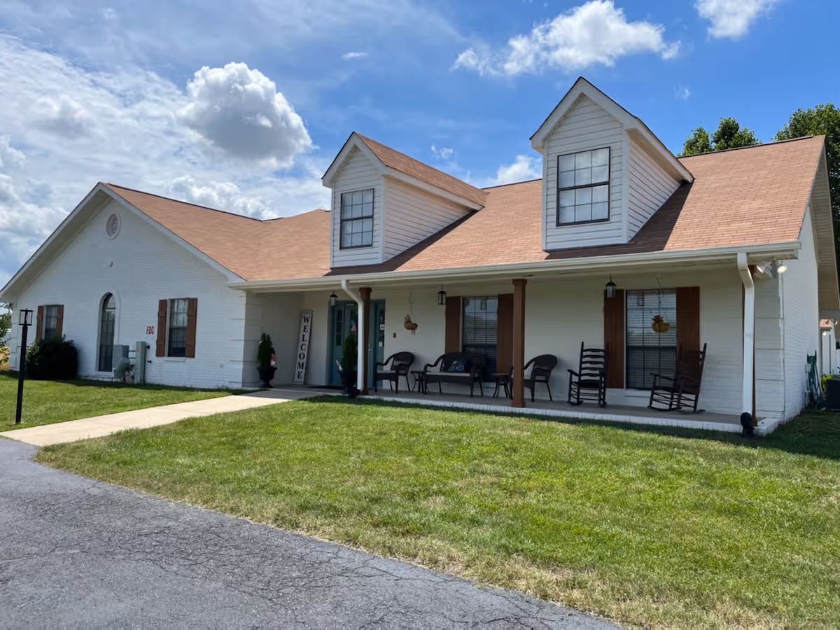 Exterior view of a single-story white brick building with a brown shingled roof and two dormer windows. The building has a covered porch with several chairs and a welcome sign near the entrance. The lawn is green and well-maintained under a partly cloudy blue sky.