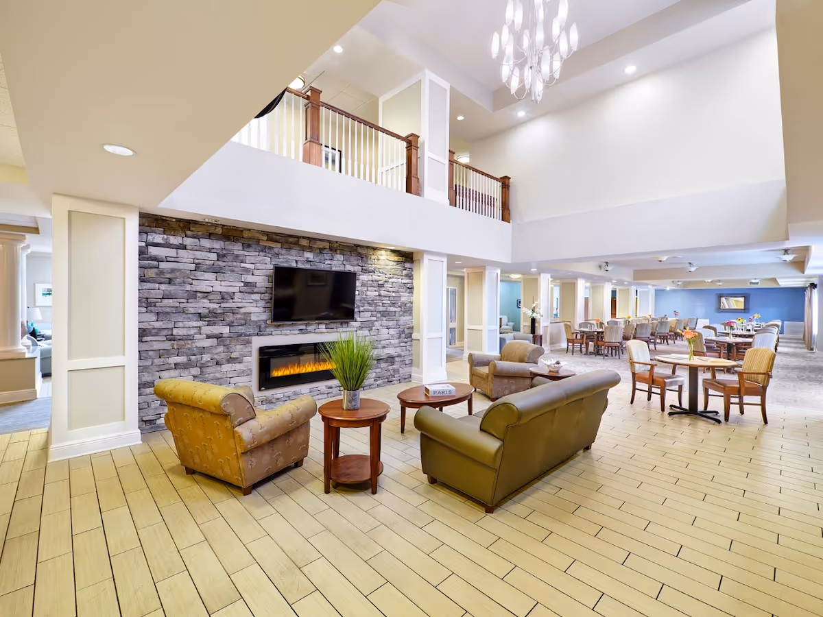 A spacious senior living facility common area with a high ceiling and a modern chandelier. The room features a stone accent wall with a mounted flat-screen TV above an electric fireplace. Comfortable seating includes a green leather sofa and two patterned armchairs arranged around wooden coffee tables with decorative items. In the background, there are multiple dining tables and chairs with flower centerpieces, set against a blue accent wall.