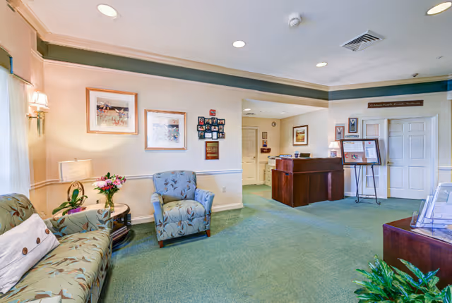 A well-lit reception area with a green carpet, floral-patterned sofa and armchair, a small round table with a flower vase, framed artwork on the walls, and a wooden reception desk in the background. There are informational displays and plaques on the walls and a potted plant in the foreground.