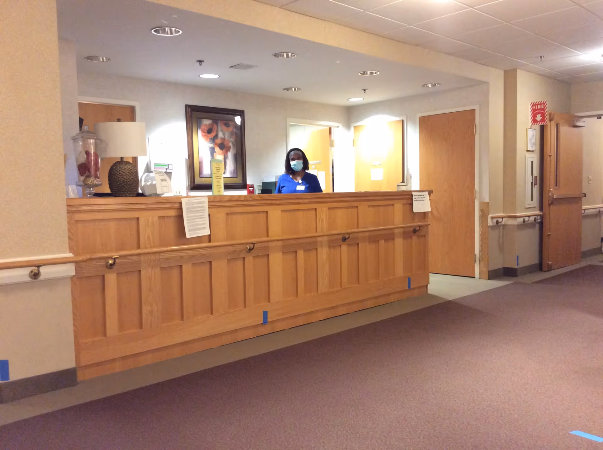 Reception area in an assisted living facility with a wooden front desk. A staff member wearing a blue uniform and a face mask stands behind the desk. The area is well-lit with ceiling lights, and there are doors and framed artwork on the walls behind the desk.