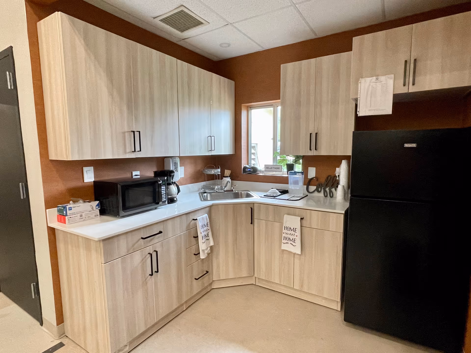 Small kitchenette with light wood cabinets, a countertop microwave and coffee maker, sink beneath a window, and a black refrigerator.
