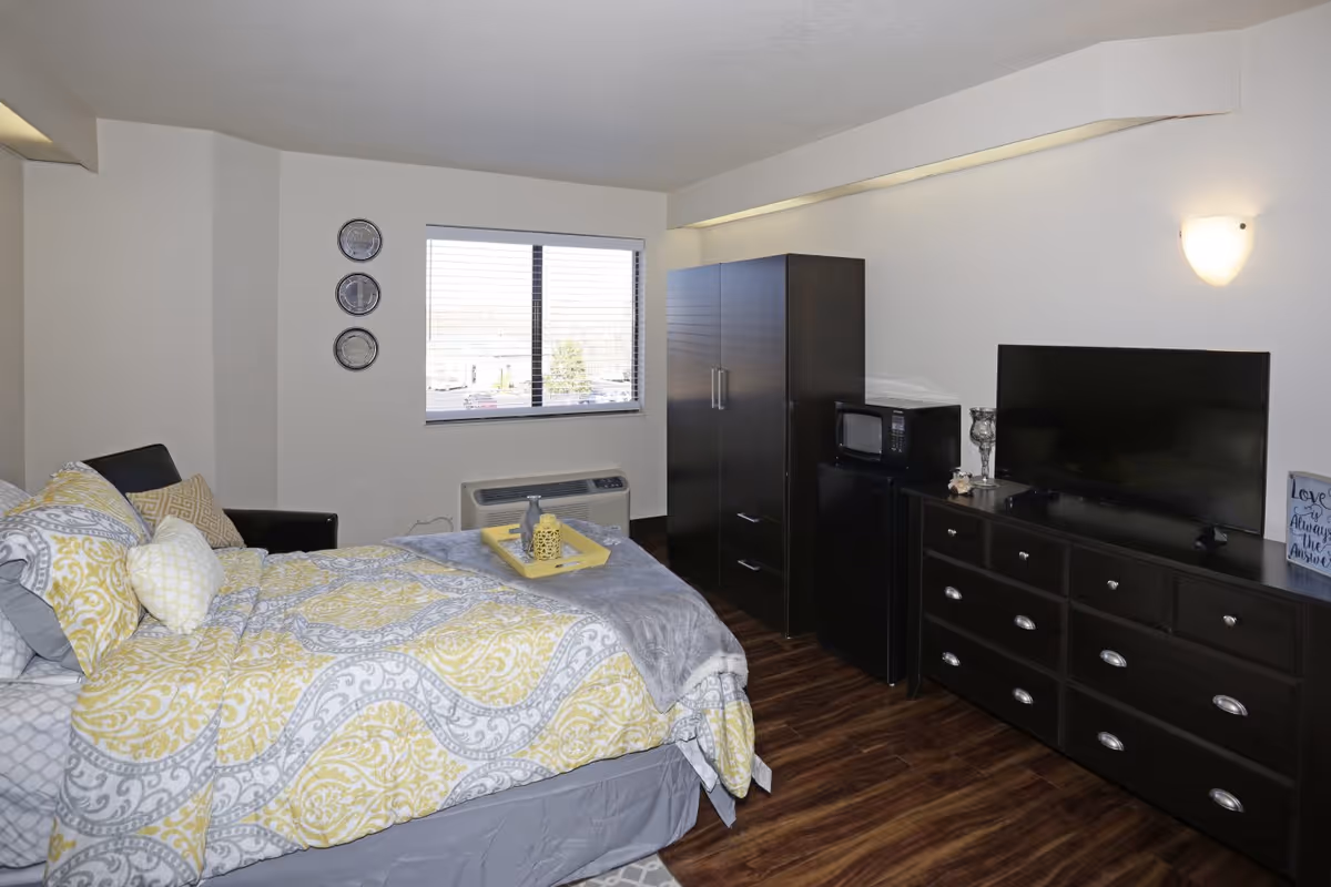 A neatly arranged bedroom with a bed covered in yellow and gray patterned bedding. There is a window with blinds, a dark wooden wardrobe, a small refrigerator with a microwave on top, and a dark wooden dresser with a flat-screen TV. The room has wooden flooring and a wall-mounted light fixture.