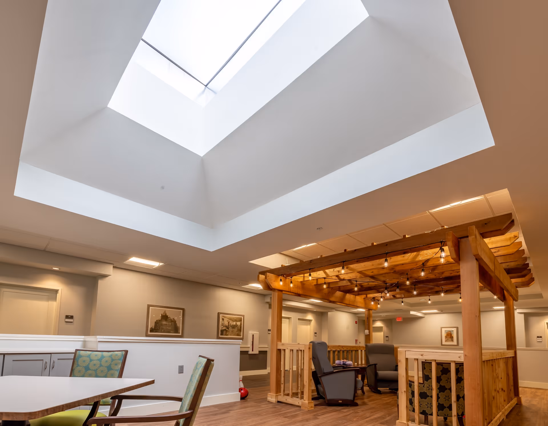 Interior view of a senior living facility common area with a large skylight ceiling, wooden pergola structure with string lights, comfortable chairs, and tables with chairs. The walls are decorated with framed pictures.