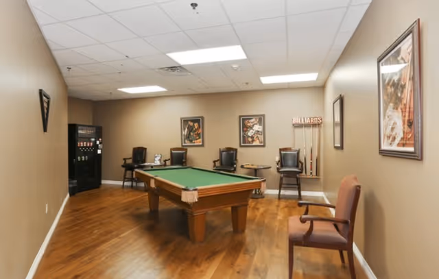 A recreational room with a pool table in the center, surrounded by several chairs along the walls. There are framed pictures on the walls, a rack holding billiard cues, and a vending machine in the corner. The room has wooden flooring and beige walls with a white ceiling featuring fluorescent lights.